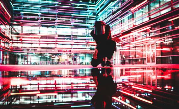 Image of a man kneeling inside a building, with neon lights and reflections, to suggest many dimensions of AI. Image by Drew Dizzy Graham, Unsplash, https://unsplash.com/license