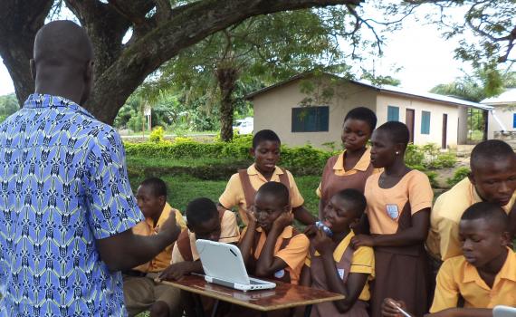 A librarian teaching a group of school children to learn computer skills. The class is taking place outside, under a tree.